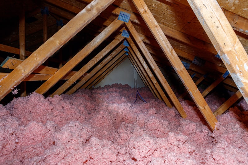 A dark attic in a wood-frame house has pink loose fill fiberglass insulation covering the floor.