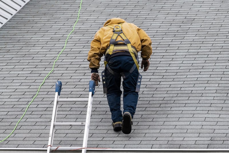 House during day with gray color Single Family Home and construction man in yellow uniform walking on roof shingles and ladder during repair