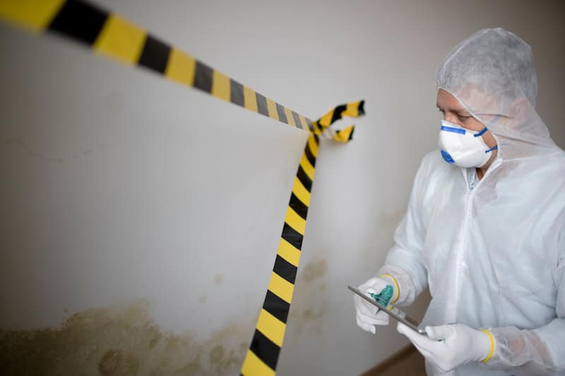 Man with white protective suit and mouth nose mask stands in front of mold on wall and works with tablet behind barrier