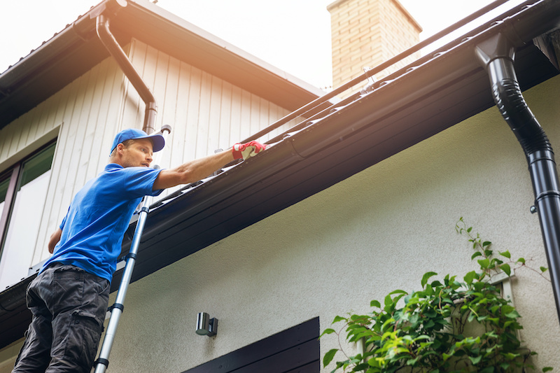 man on ladder cleaning house gutter from leaves and dirt drainage