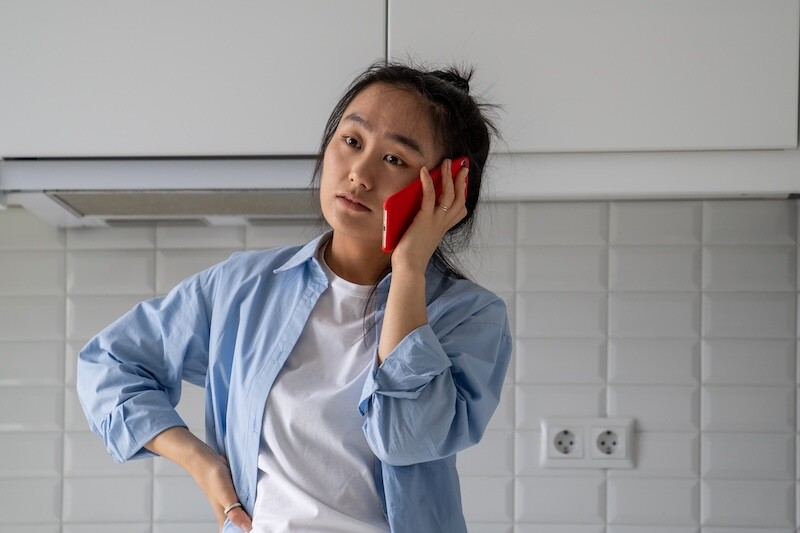 Upset dissatisfied Asian woman talking on phone arguing by mobile while standing in kitchen at home, talking about problems with friend. Unhappy girl waiting on hold, calling customer service