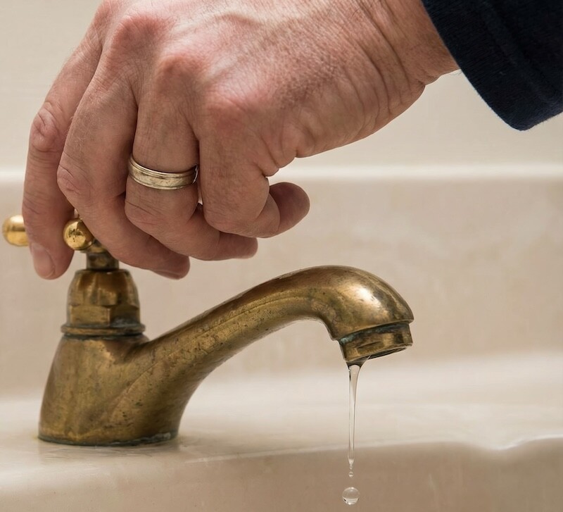 A hand turns a faucet in a bathroom. Water drips from the spout. There is a note on the wall that says every drop counts. The setting emphasizes water conservation.