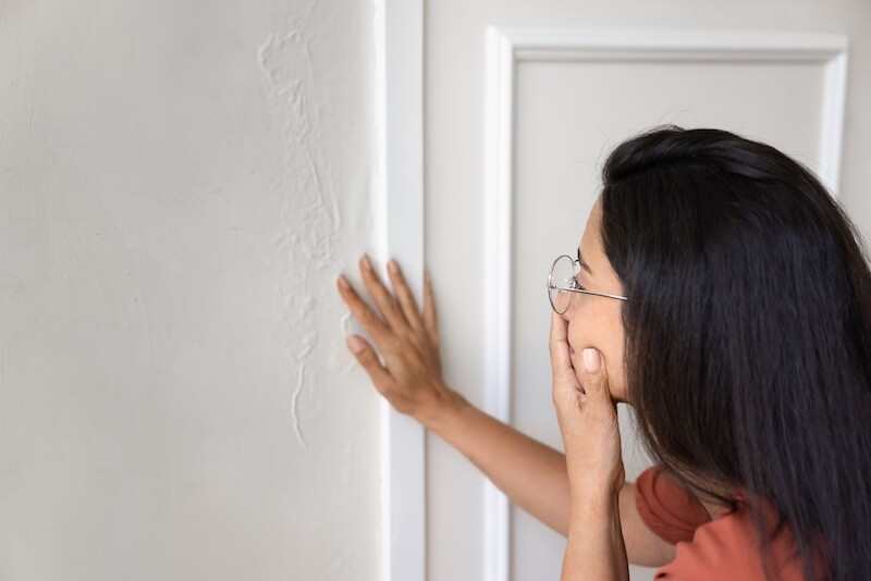 Close-up view of concerned, shocked young woman wearing glasses touching damaged interior wall with visible cracks or water stains. Home maintenance issues, property inspection, and renovation needs