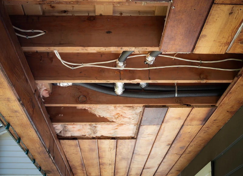 Exposed insulation and wrapped plumbing pipes in open ceiling cavity. Discolored fiberglass batt with gaps and foil wrapped drain pipes for freeze protection. Selective focus.