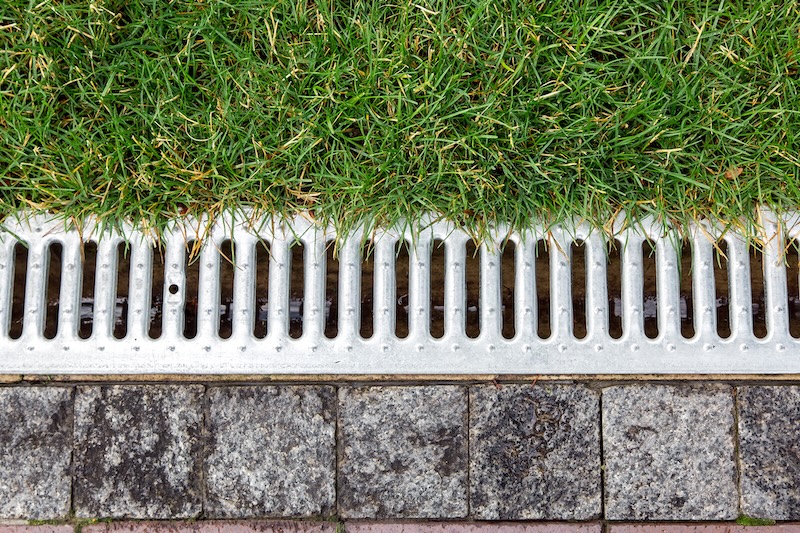 iron grate of a storm drainage system on the side of a footpath made of pavers near a green lawn top view.