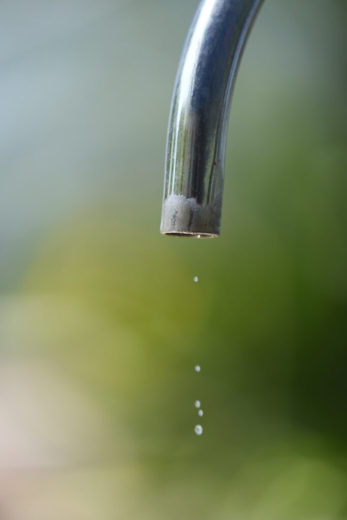 Water faucet droplet metal pipe spout splash drip, closeup of stainless steel tap releasing small water drops against green bokeh background, concept of leak and water saving