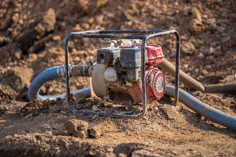 Portable dewatering pump with blue suction hoses on rough excavation ground for trench drainage, water removal, and construction site utility work